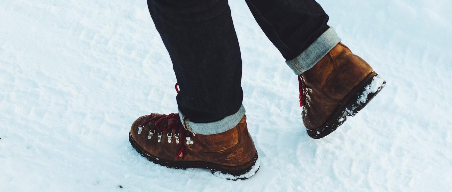 Personne portant des chaussures de randonnée marron à lacets rouges et un jean foncé retroussé, marchant dans la neige.