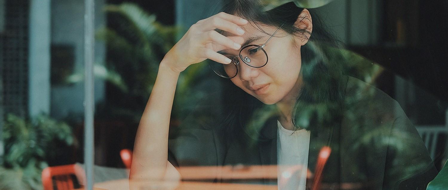 Personne à lunettes à l'air stressé, main sur le front, assise près d'une fenêtre avec des reflets de plantes dans des tons verts atténués.