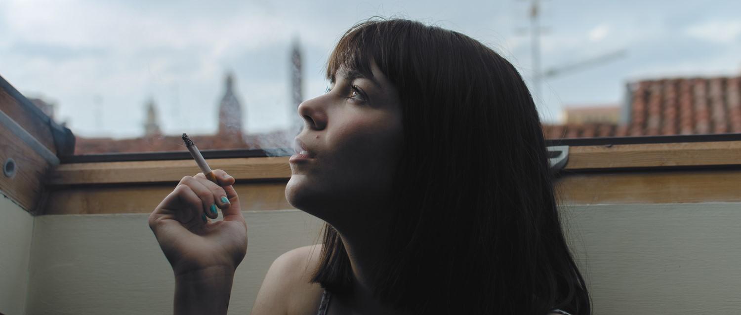 Personne aux cheveux noirs fumant près d'une fenêtre, regardant pensivement l'horizon de la ville, vernis à ongles sarcelle visible.