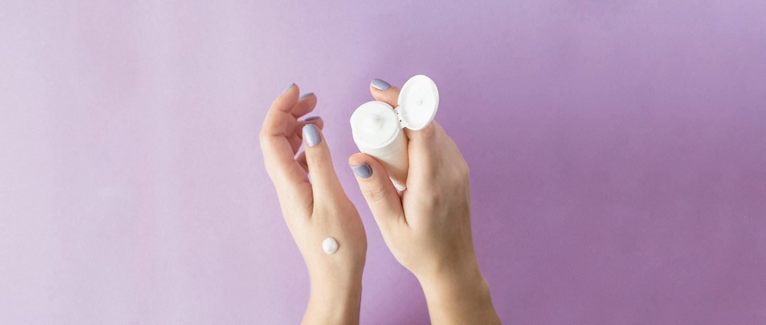 Mains avec vernis à ongles bleu pâle tenant d'une main un récipient de soin blanc avec de la crème sur un fond violet.
