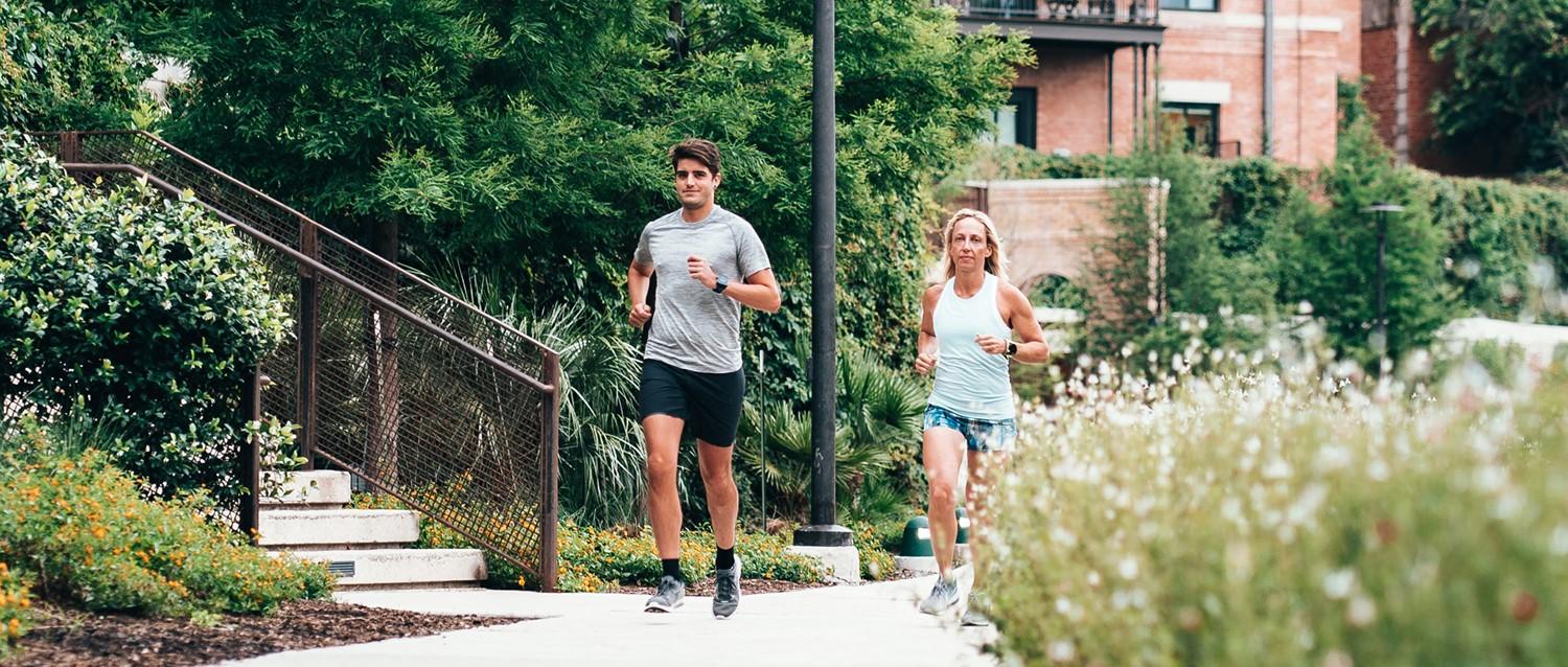Deux coureurs font leur jogging le long d'un sentier de jardin avec de la verdure luxuriante et des bâtiments en briques en arrière-plan.