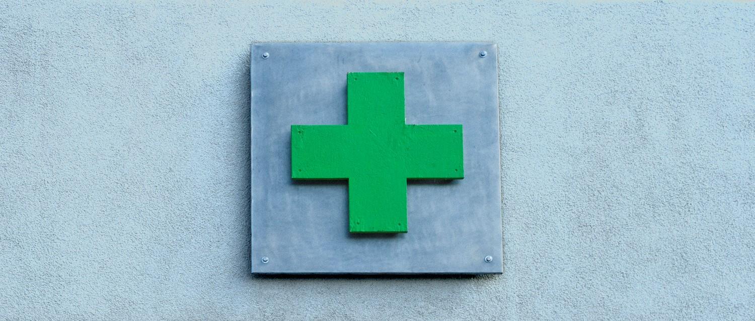 Croix médicale verte montée sur une plaque métallique grise contre un mur bleu clair, indiquant un établissement de soins de santé.