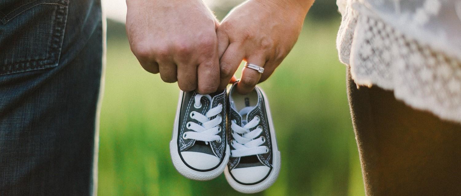 Couple se tenant par la main et portant de petites chaussures de bébé noires, l'une des personnes portant une bague de fiançailles sur fond vert.