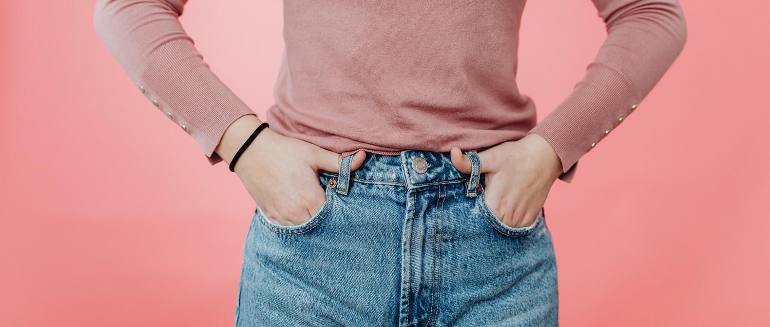 Personne portant un pull rose avec les mains dans les poches d'un jean bleu sur fond rose corail.